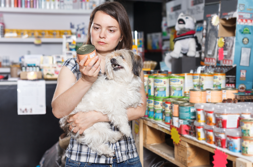 Portrait of woman with small dog choosing preserves in pet shop