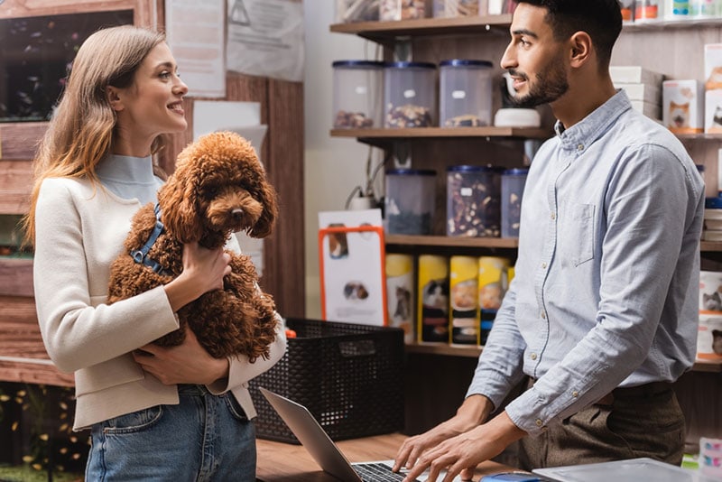 smiling woman holding poodle dog and talking to a man in pet shop